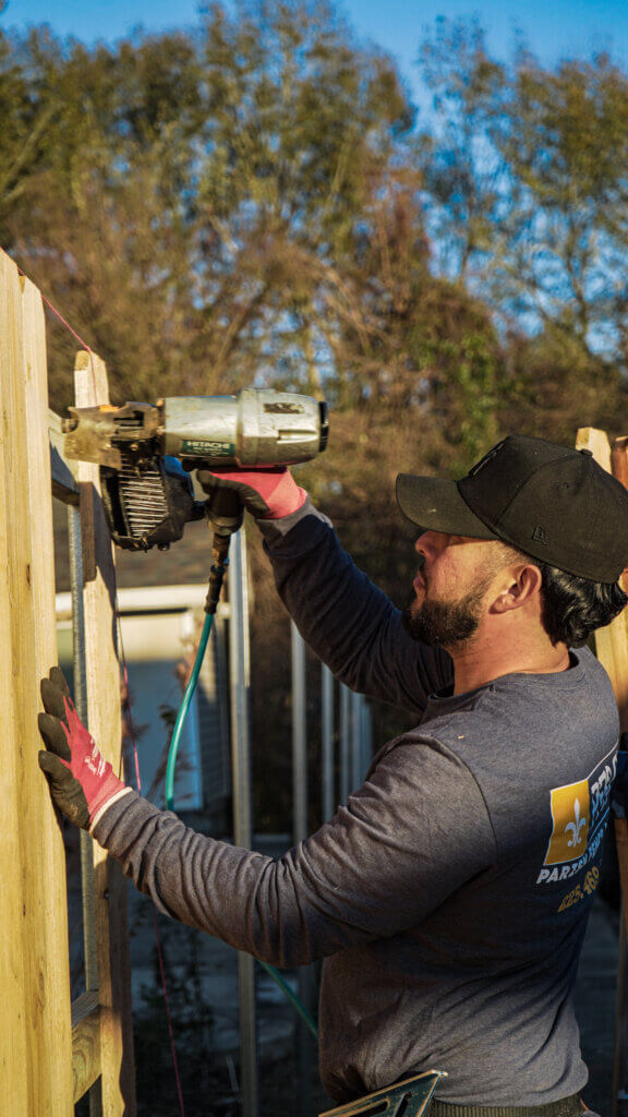 A man wearing a black cap and gloves uses a nail gun to secure wooden boards while constructing a fence outdoors for a Fence Company in Baton Rouge, LA. Trees and blue sky can be seen in the background.