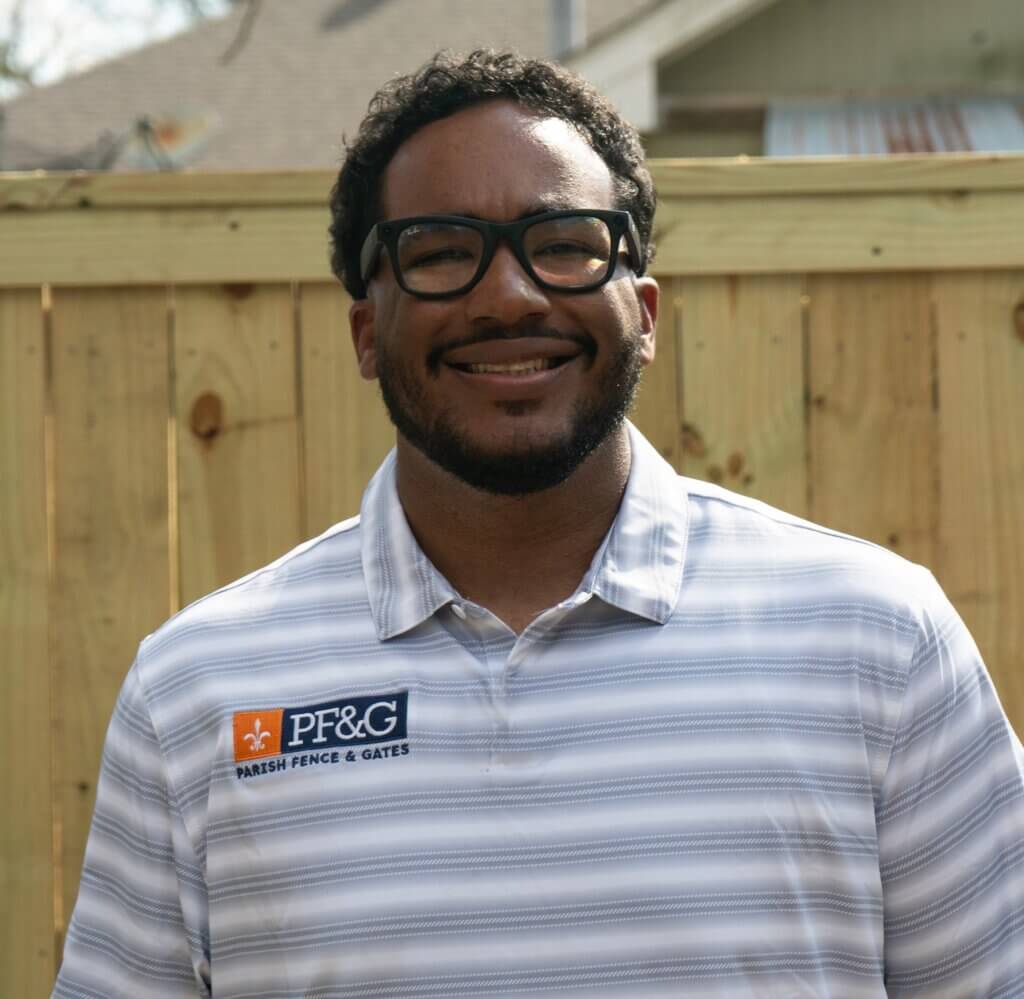 A man with short dark hair, beard, and glasses is smiling while standing outside in front of a wooden fence. He is wearing a striped polo shirt with a "PF&G Parish Fence & Gates" logo, representing a trusted Fence Company in Baton Rouge, LA.